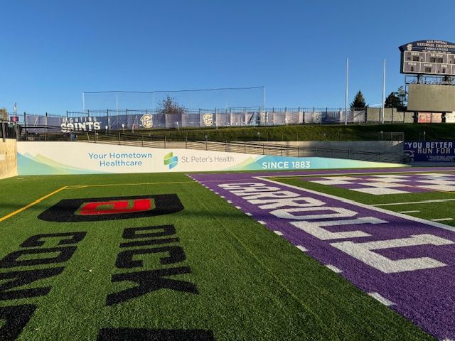 Football field with sponsor sign, purple and green turf, and a scoreboard under a clear blue sky.