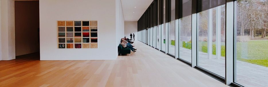 An art museum interior with a wood floor, rectangular art pieces on the wall, and large windows overlooking a grassy outdoor area.