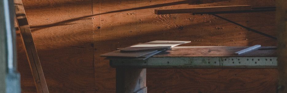 A dimly lit workshop with a wooden table, tools, and a ladder. Sunlight streams across the wall.