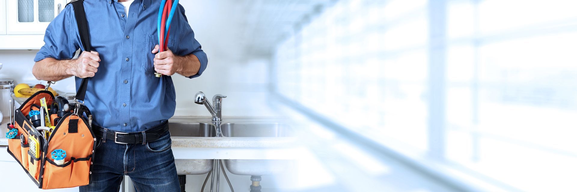 Plumber holding pipes with a tool bag beside a kitchen sink.