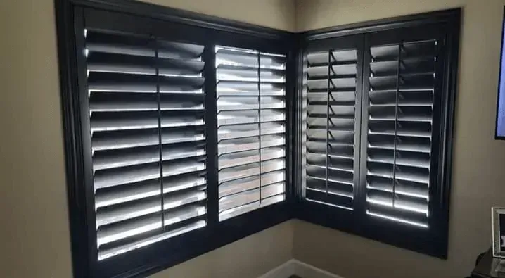 Corner window with black shutters, angled and partially open, in a room with beige walls.