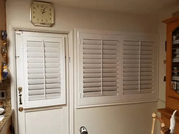 White shutters cover a door and a window in a kitchen, a clock hangs above the door.