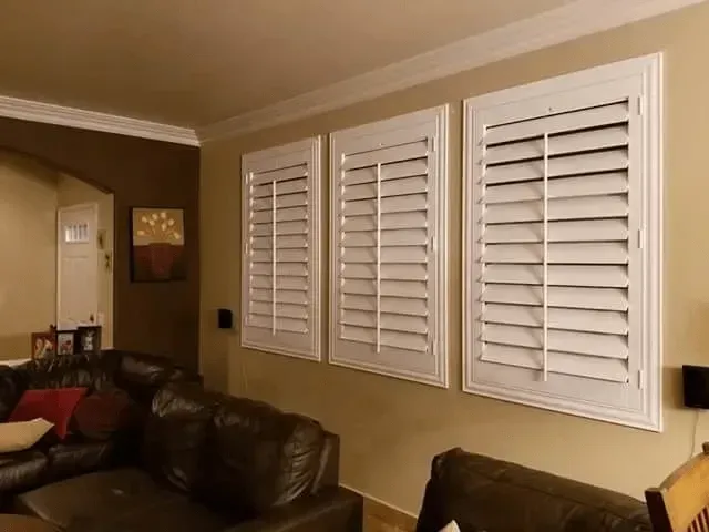 Three white shutters on a beige wall above a brown leather sofa in a living room.