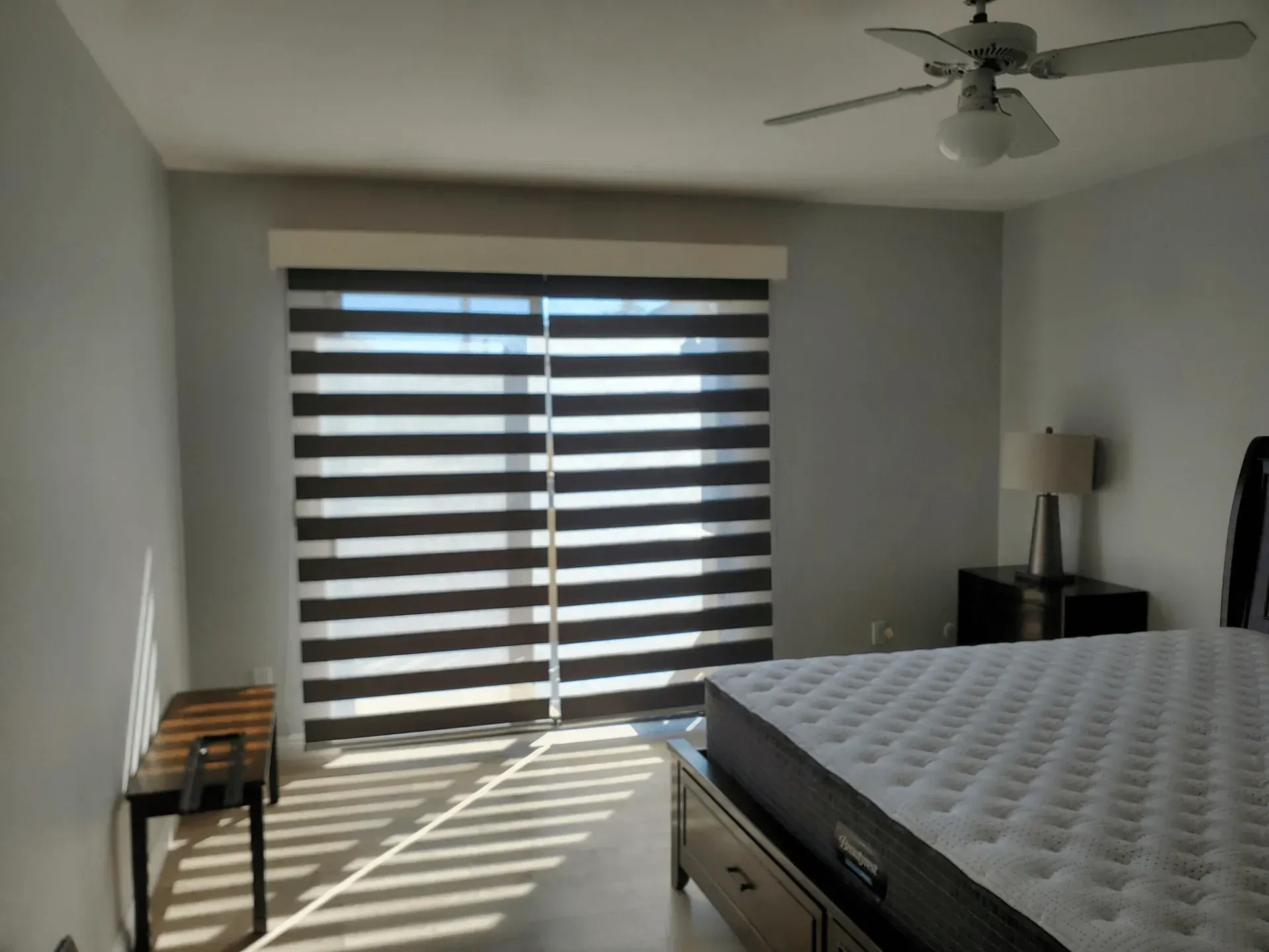 Bedroom with horizontal zebra blinds, bed, nightstand with lamp, and a bench. Sunbeams on the floor.