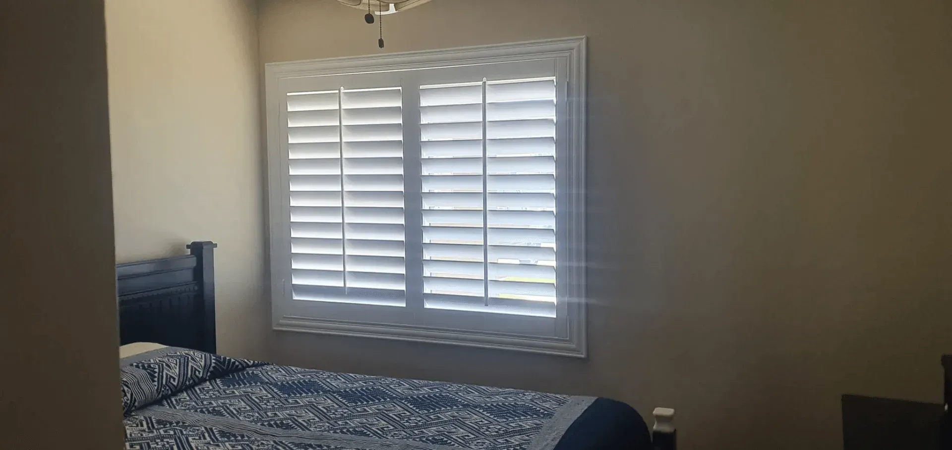 Bedroom with white shutters on the window and a bed with a blue patterned comforter.