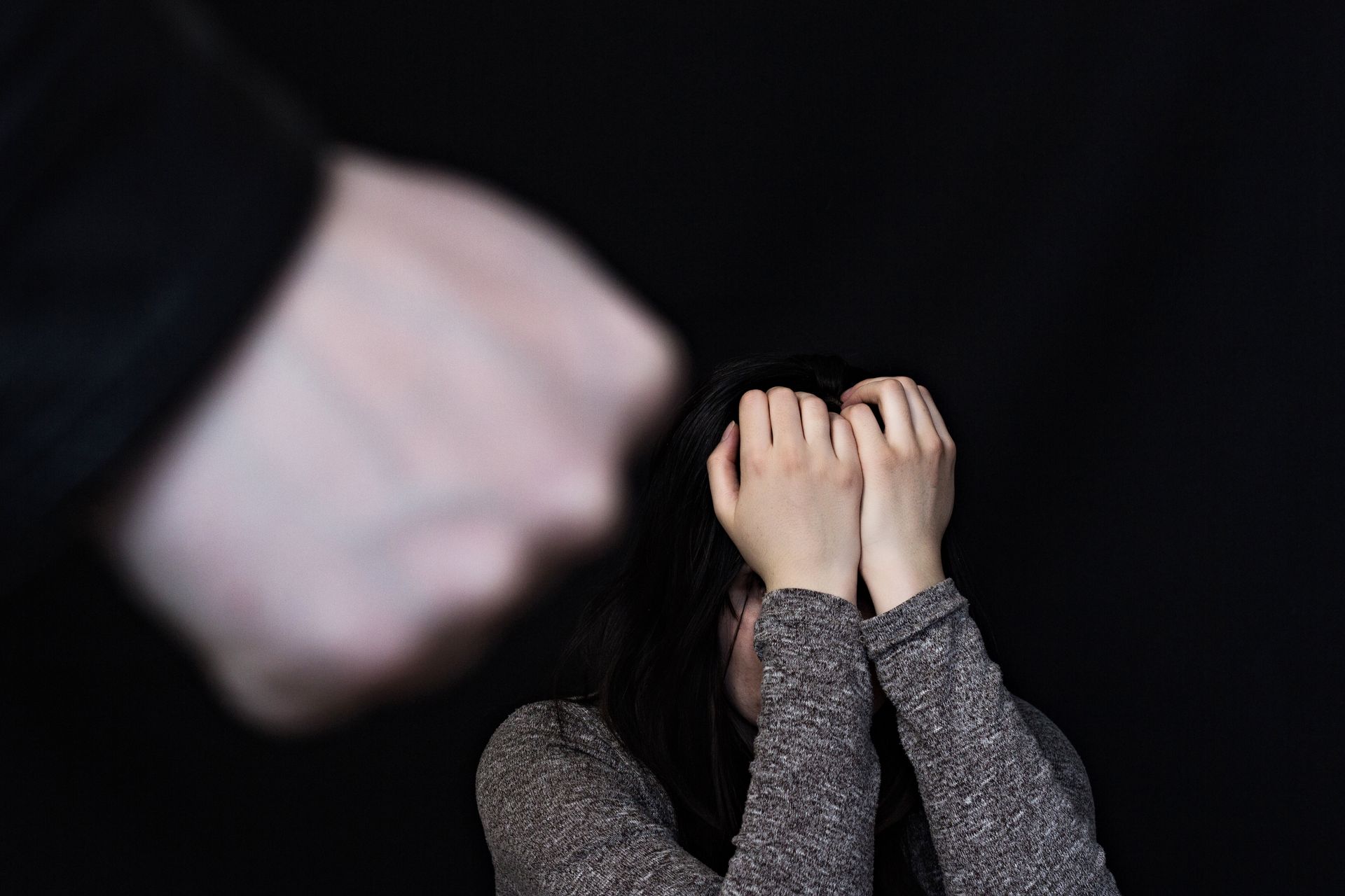 A person with hands over face cowers as a fist approaches against a black background.