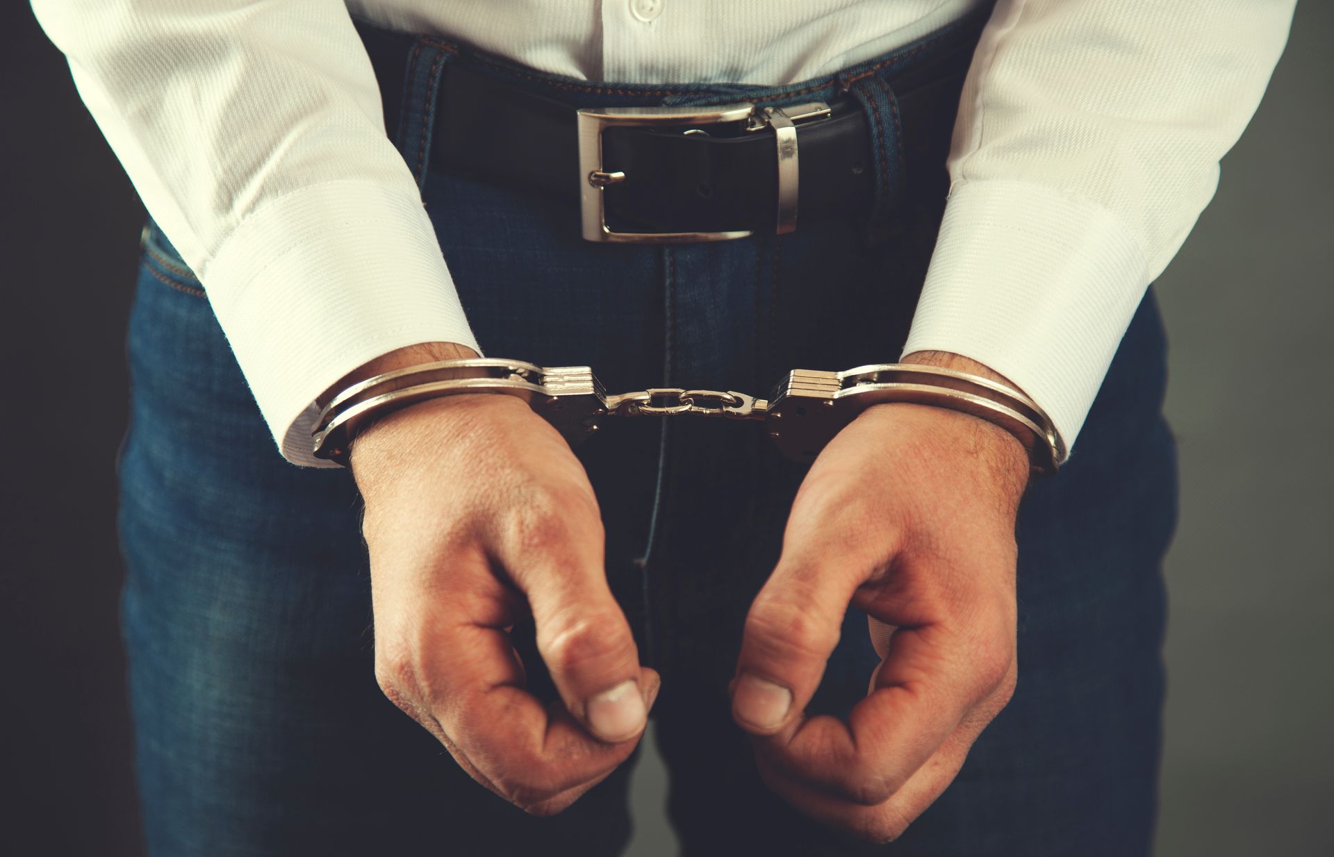 Person's hands cuffed, wearing a white shirt and jeans; visible belt buckle.