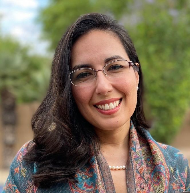 Smiling person with glasses and long dark hair, wearing a patterned shawl outdoors with blurred greenery behind