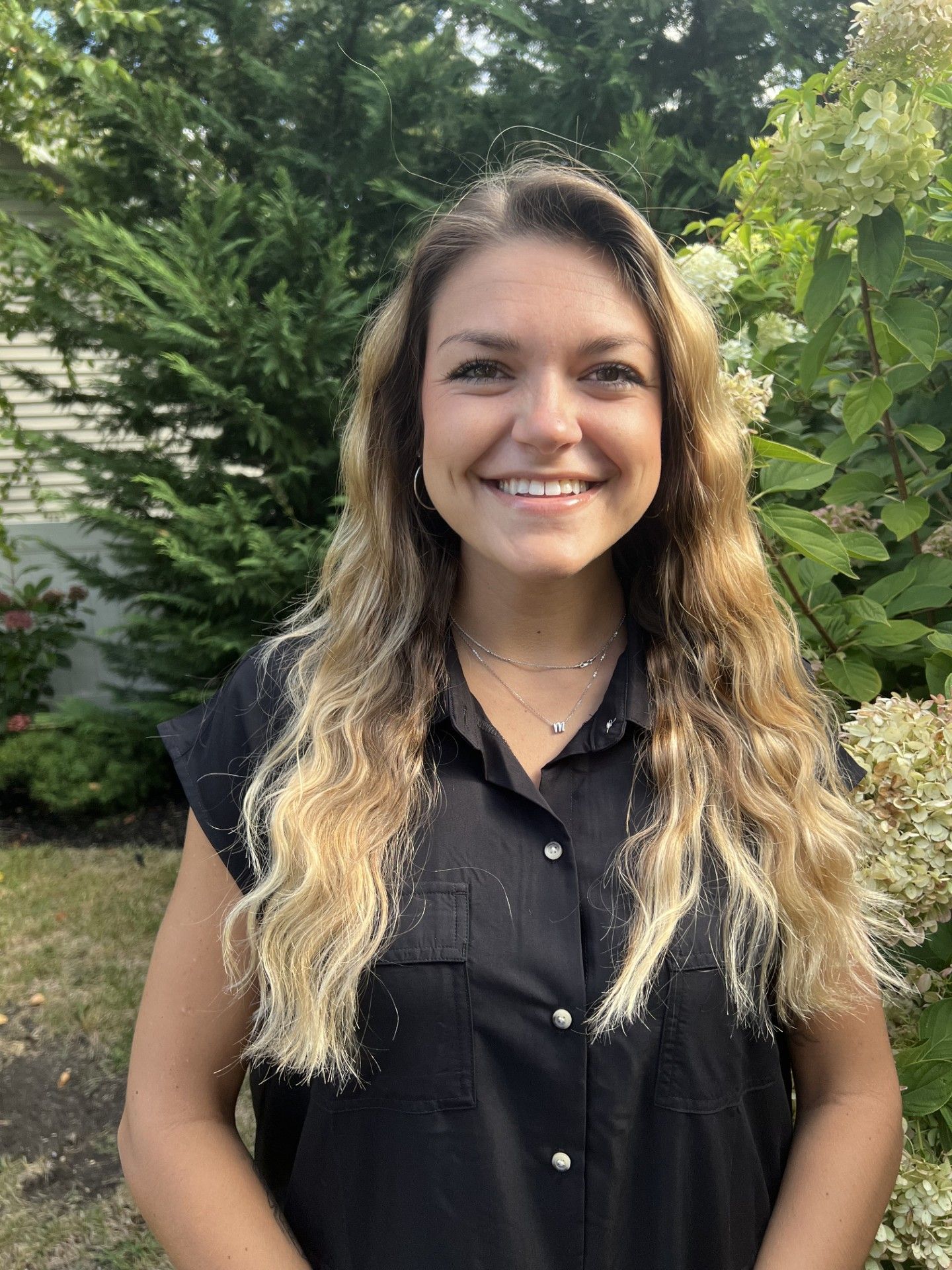 Smiling person standing outdoors in a black shirt with greenery in the background