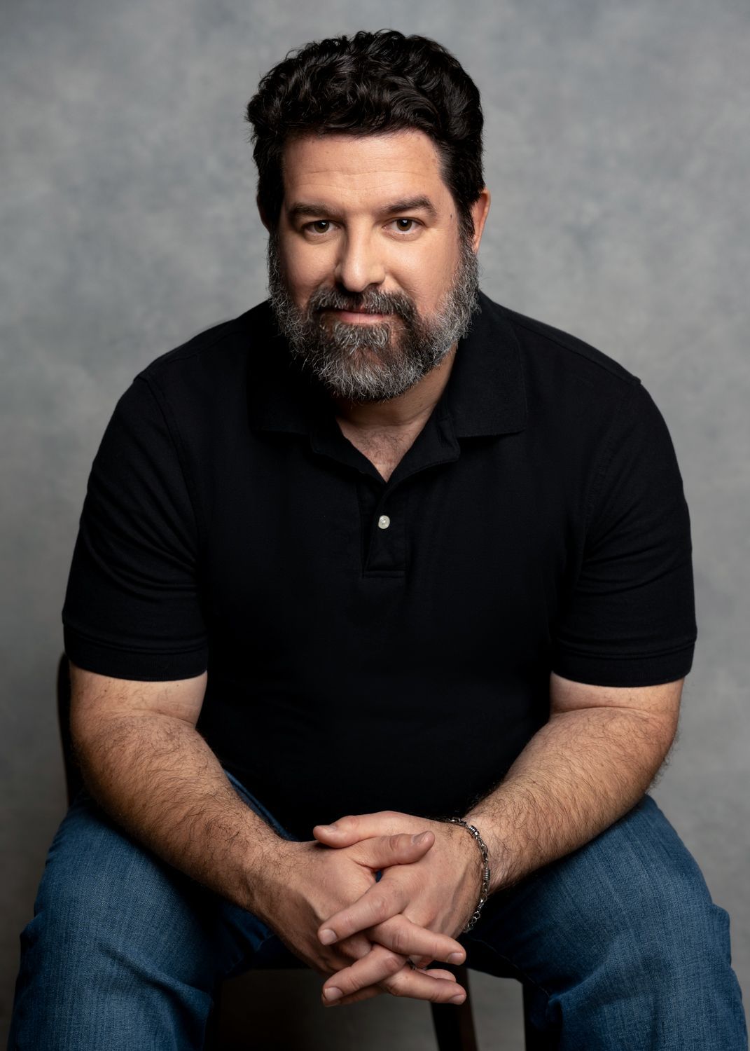 Portrait of a seated man in a black polo and jeans, hands clasped, against a gray backdrop