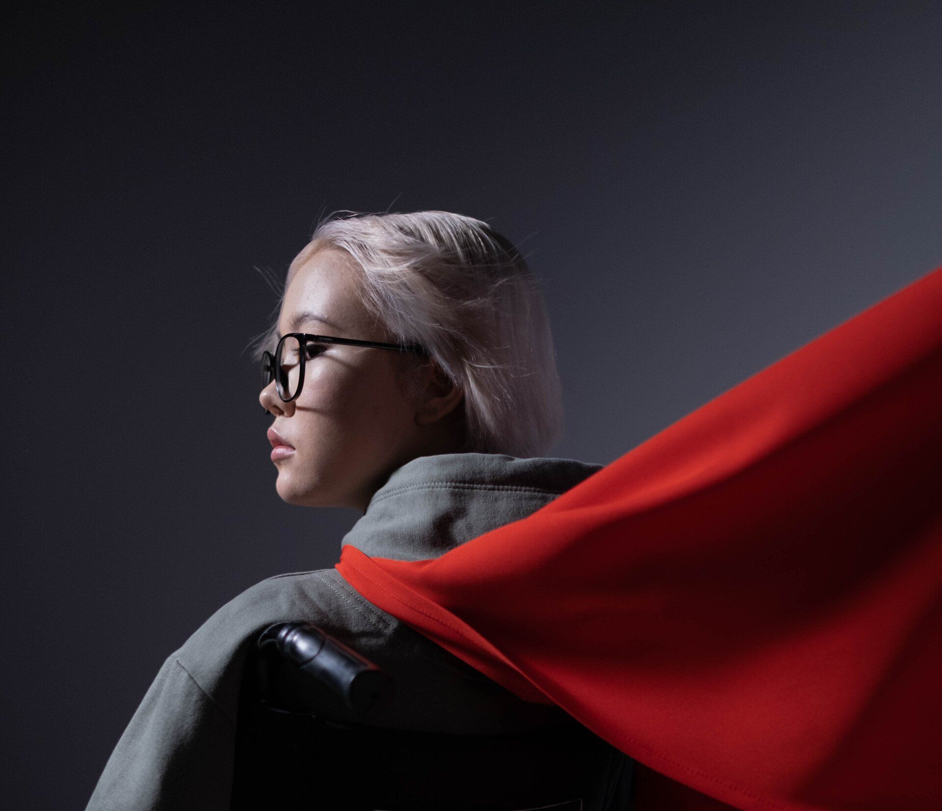 Profile of a person with glasses and light hair, wearing a gray hoodie and a flowing red cape on a dark background