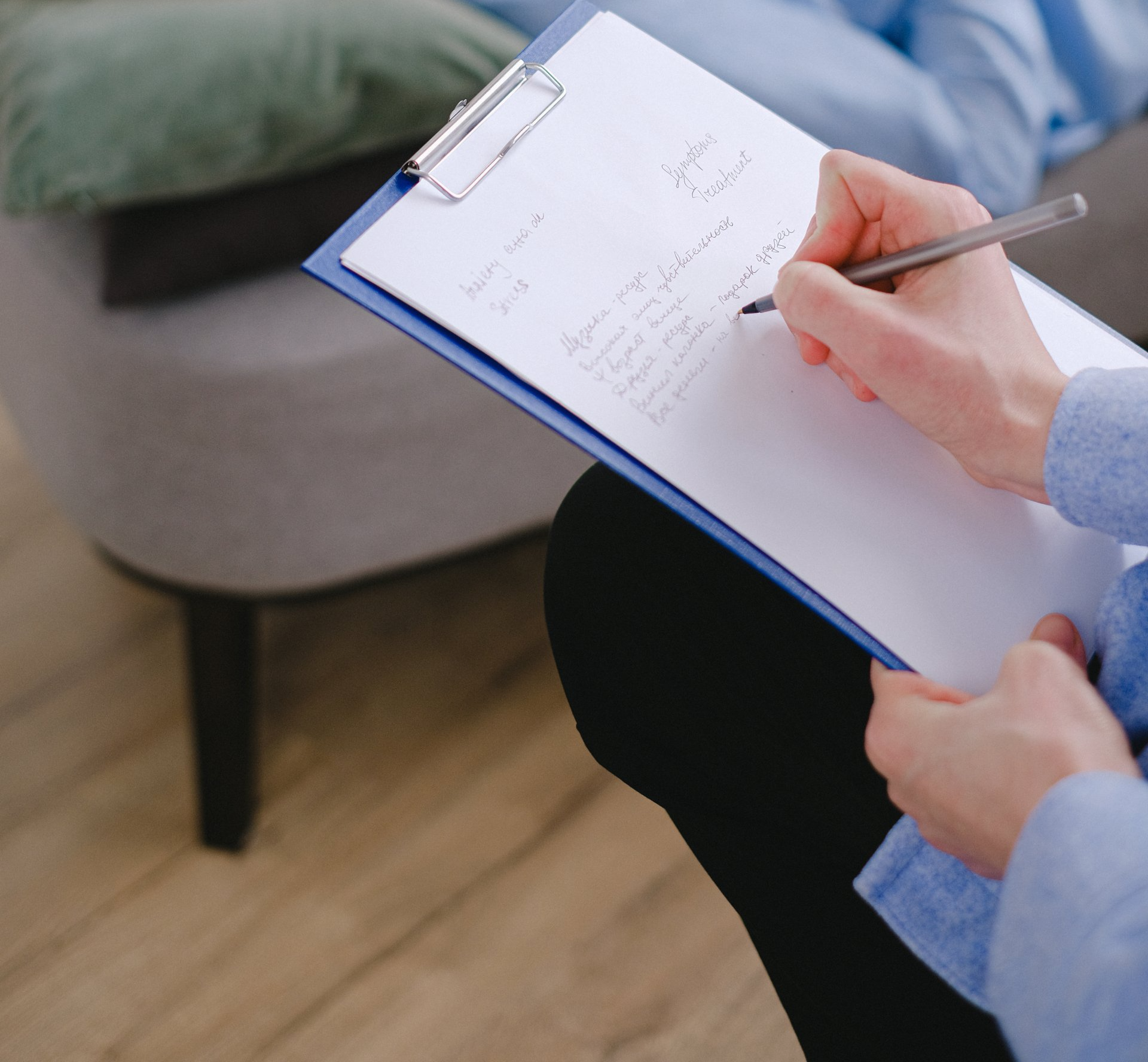 Person writing on a clipboard while seated near a gray chair and wooden floor