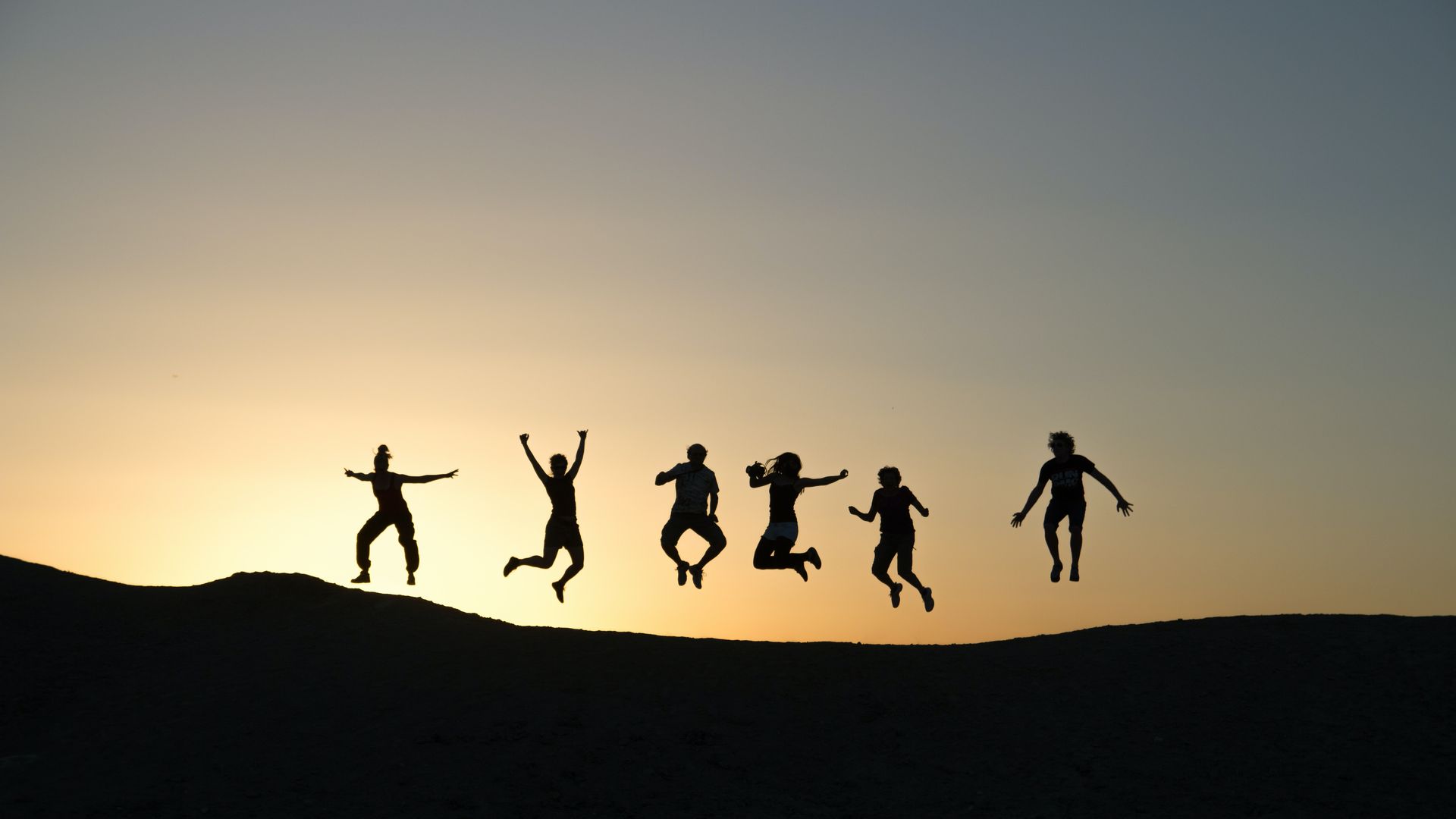Silhouettes of six people jumping on a hill at sunset under a golden sky