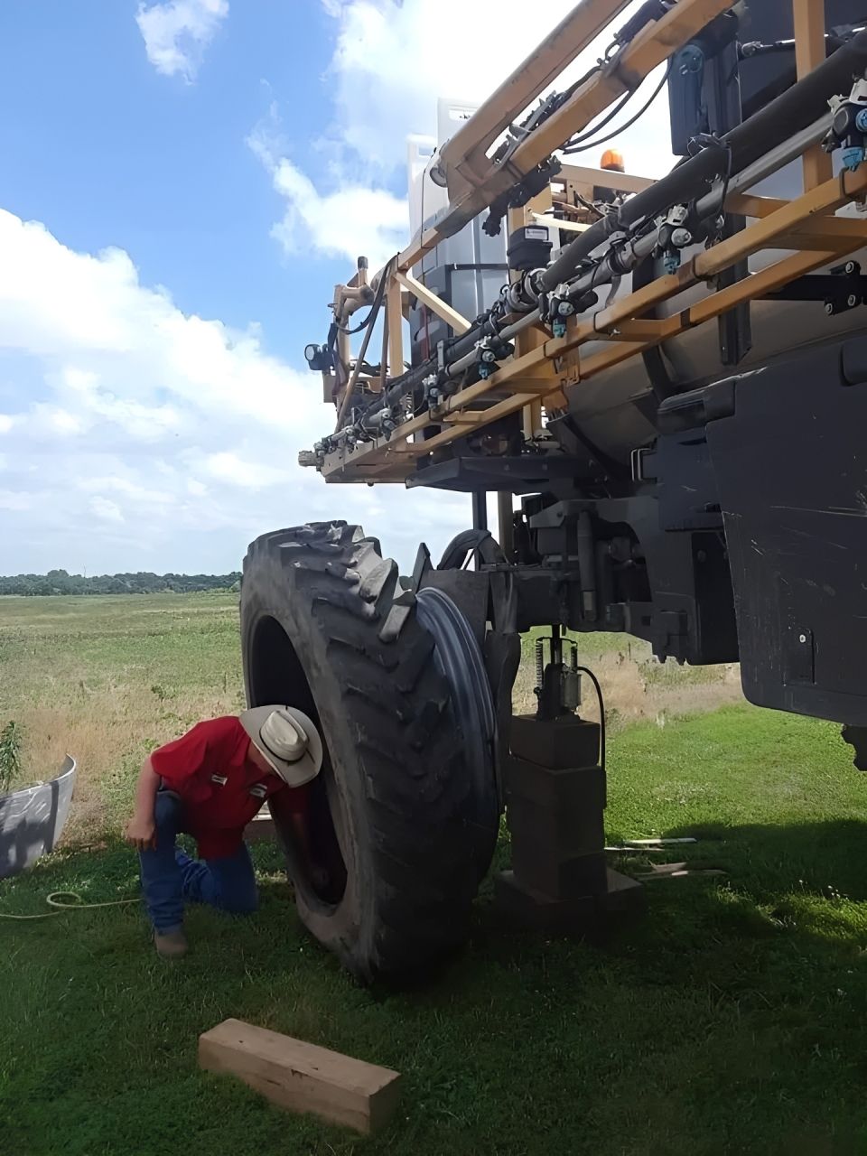 Worker in red jacket inspecting a large farm sprayer tire in a grassy field under a blue sky