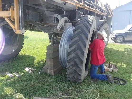 Man changing a large tire on a sprayer. The vehicle is lifted on blocks in a grassy area.