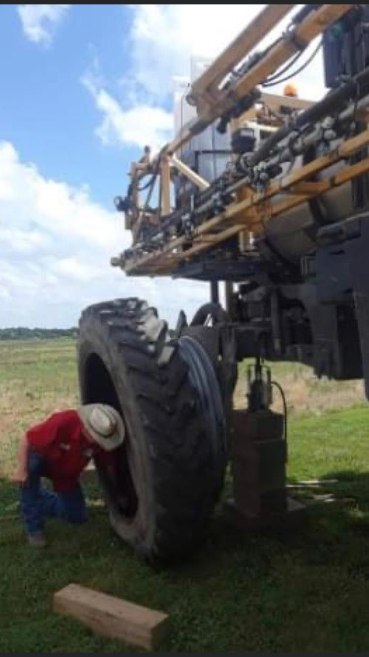 A person in a red shirt and cowboy hat works on a large agricultural sprayer's tire outdoors.