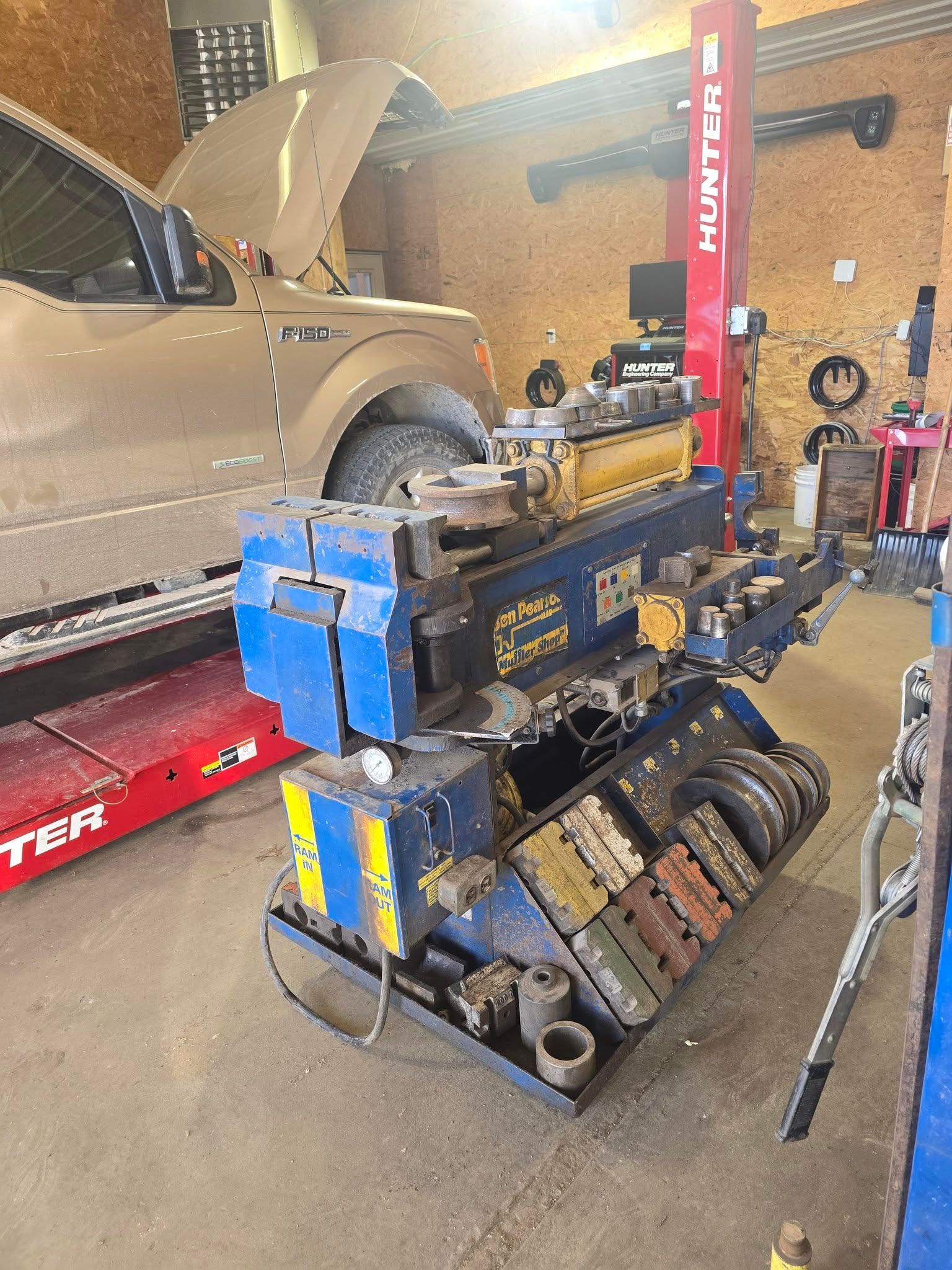 A blue and yellow exhaust pipe bender in an auto repair shop with a vehicle in the background.