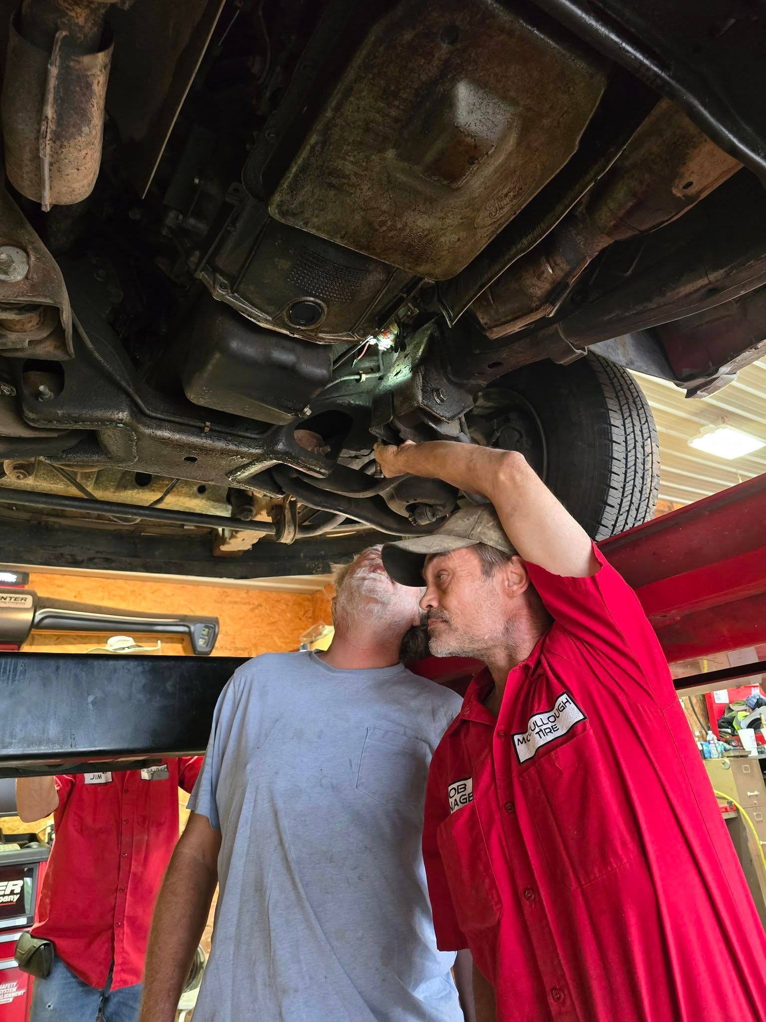 Two men under a car on a lift; one in red, leaning up; other in gray, looking up.