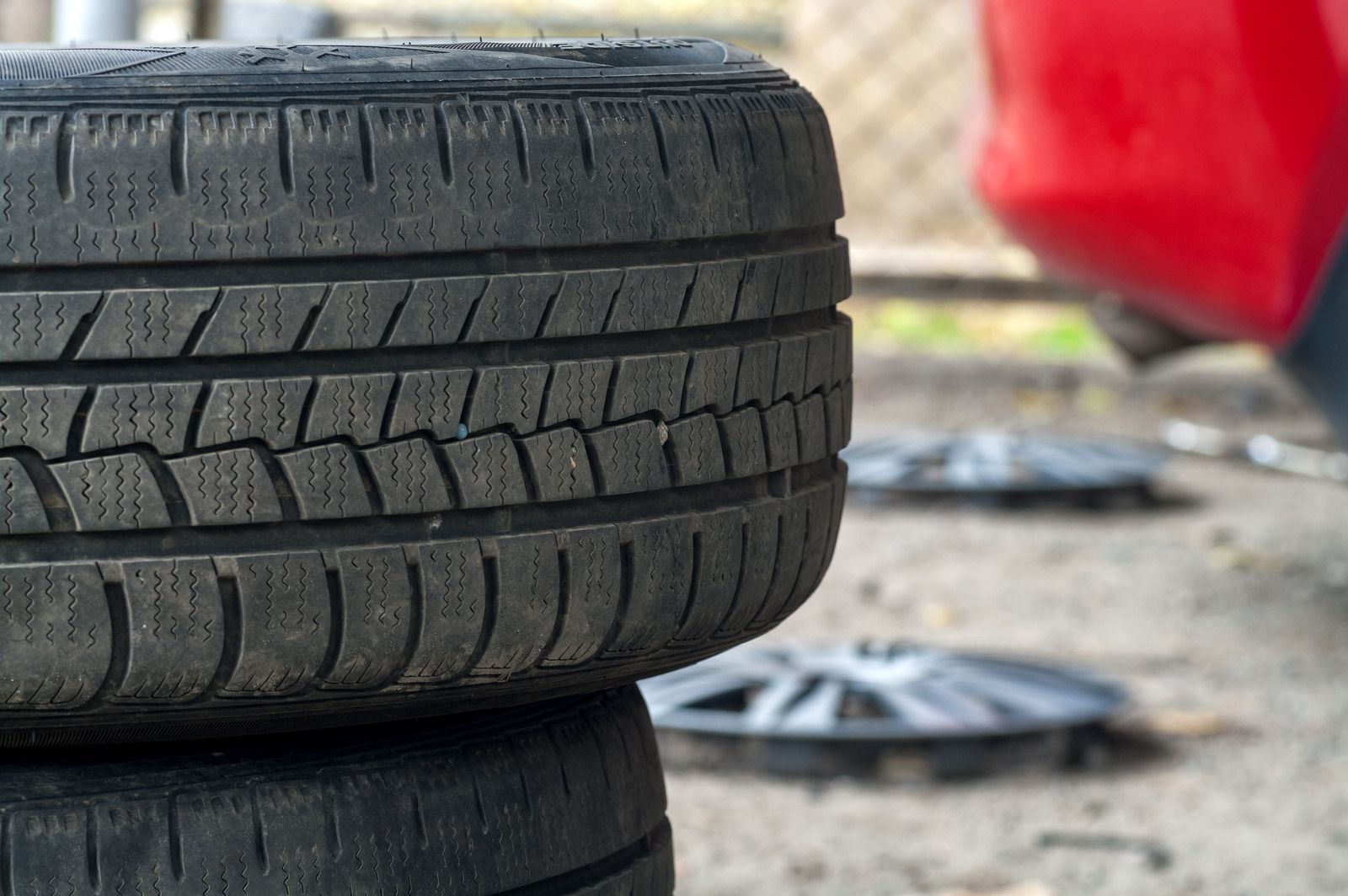Stacked car tires beside a red vehicle on a gravel surface