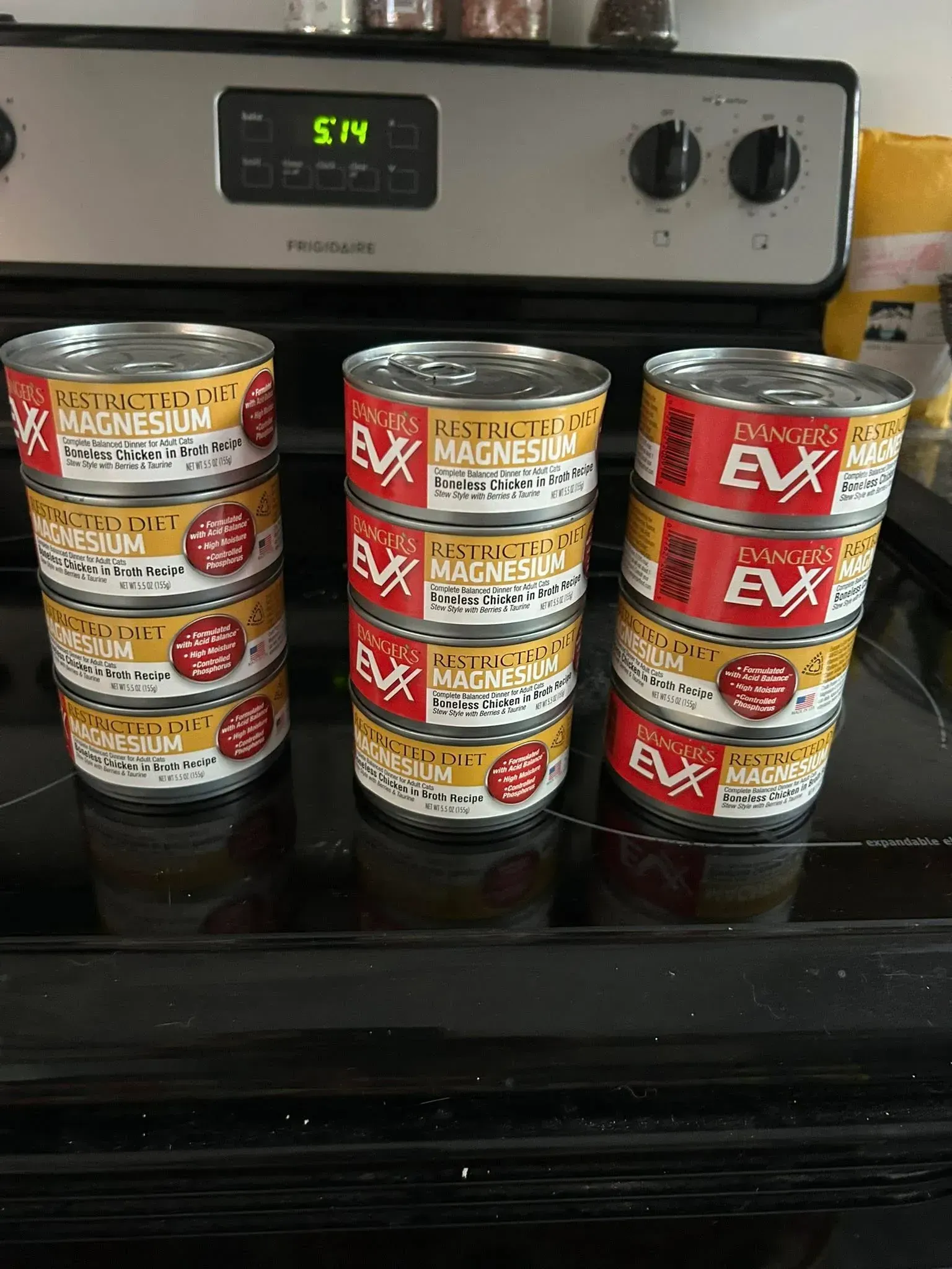 Stacked cans of cat food on a stove beneath a digital clock.