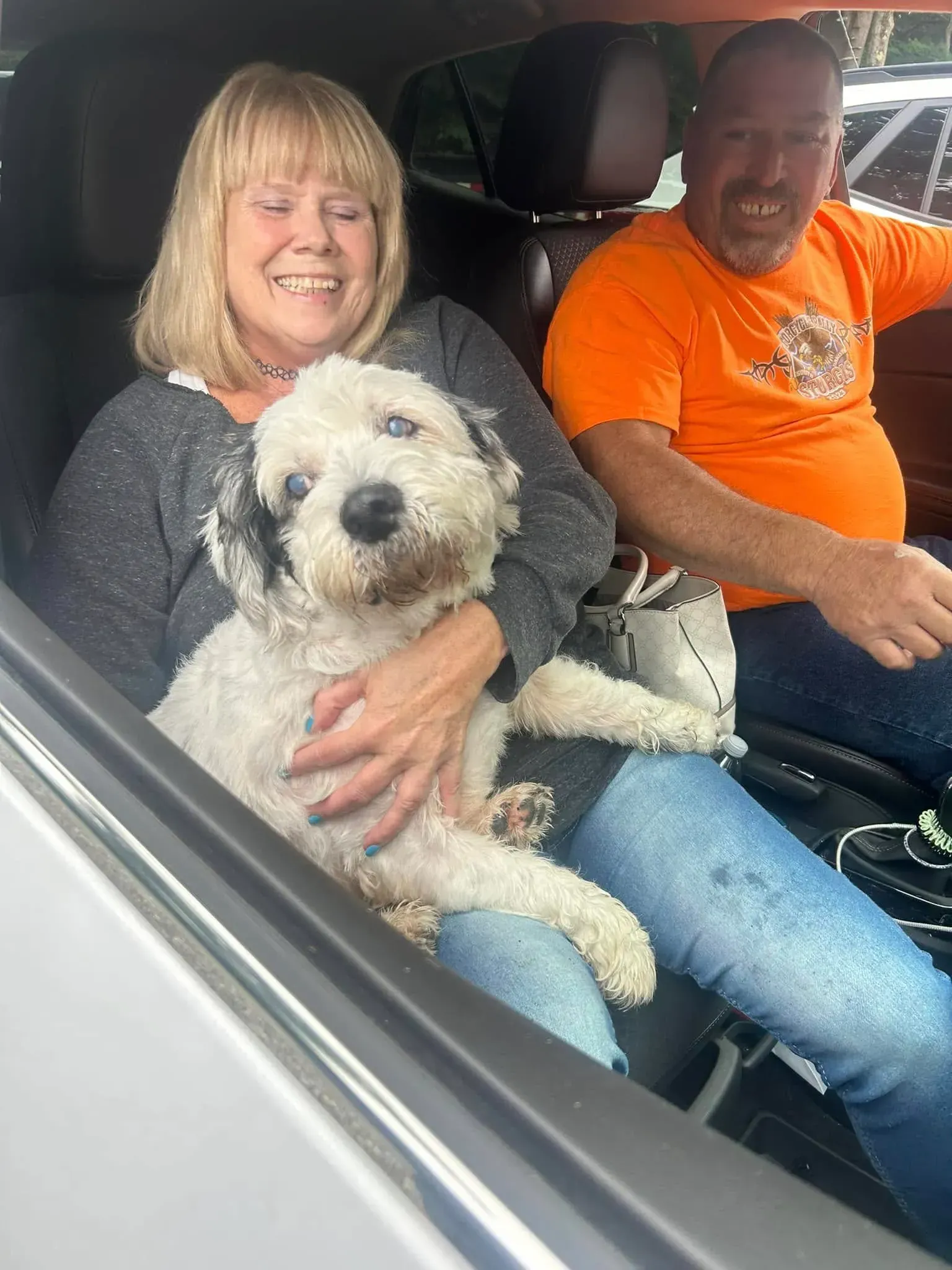 Smiling woman holding a fluffy white dog in a car, with a man in an orange shirt beside her.