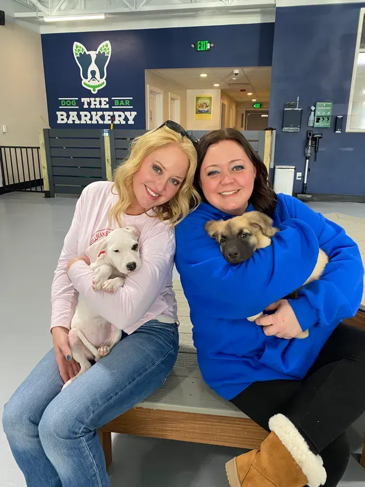 Two women sit with small dogs in a pet grooming shop called The Barkery.