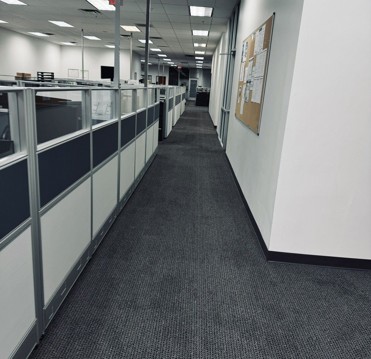 Empty office hallway with gray carpet, cubicle partitions, and a bulletin board on the wall