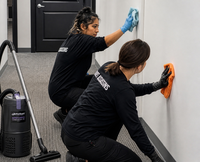 Two cleaners wiping a wall in a hallway, one with a blue cloth and one with an orange cloth.