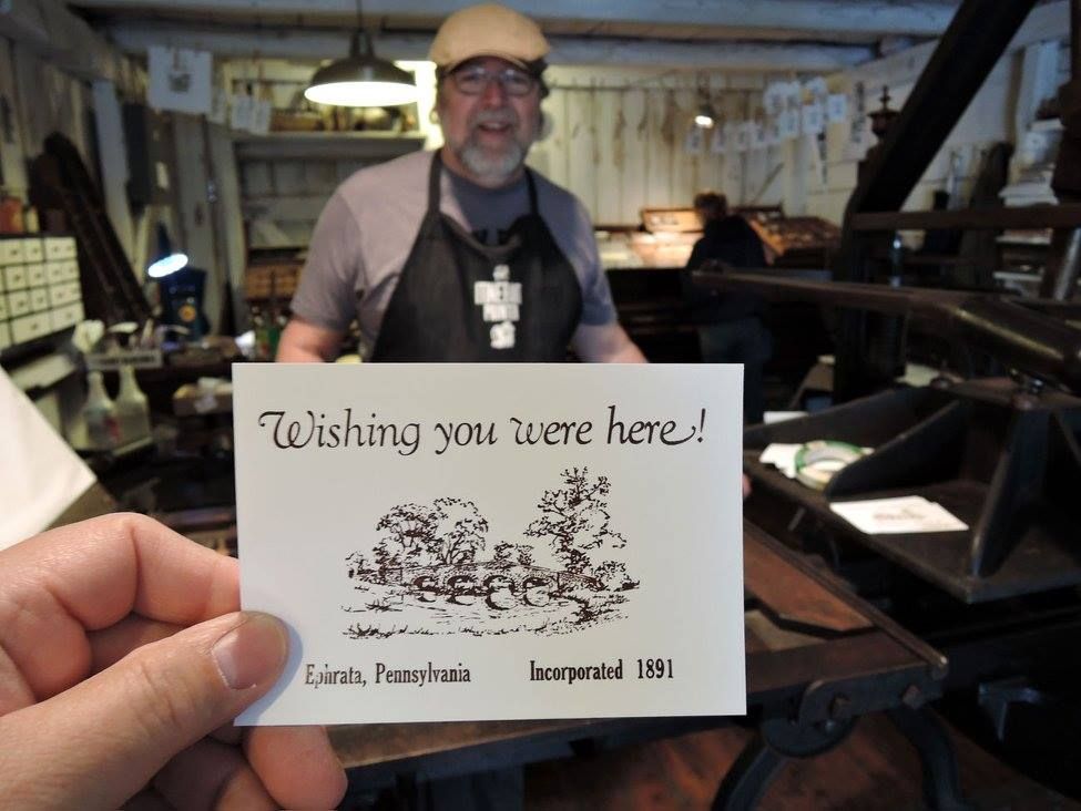 Hand holding a “Wishing you were here!” card in front of a man smiling in a workshop
