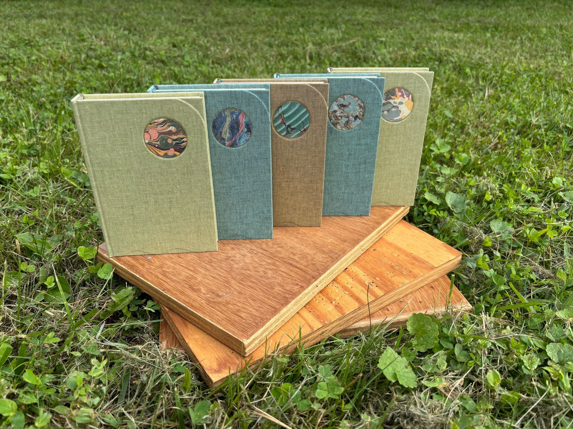 Five decorative notebooks with textured covers stacked on wooden boards outdoors on grass