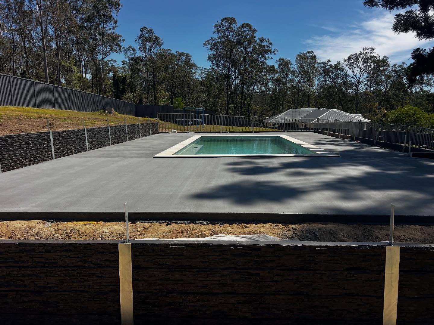 Modern backyard with a rectangular pool, large concrete deck, and trees under a clear blue sky