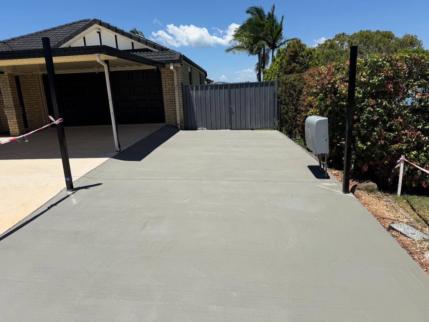 Freshly paved driveway leading to a garage, with a gray fence and tropical plants in the background