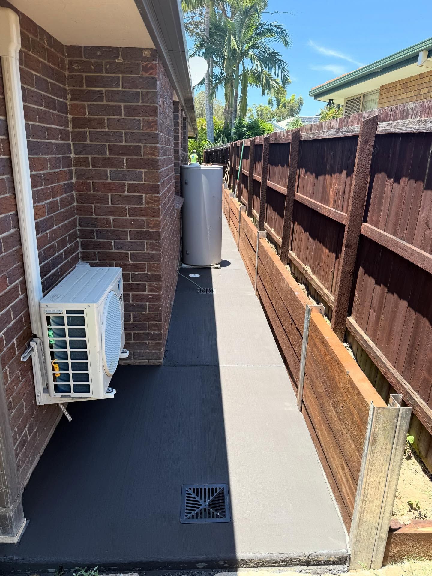 Narrow side yard beside brick house with concrete path, air conditioner units, wooden fence, and palm trees beyond.