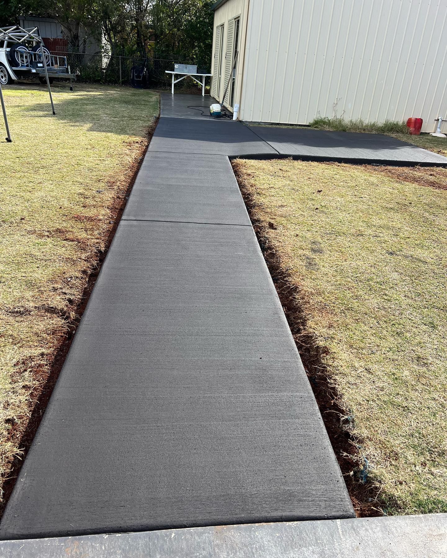 Concrete sidewalk along a house beside a grassy yard, leading to a small patio area.