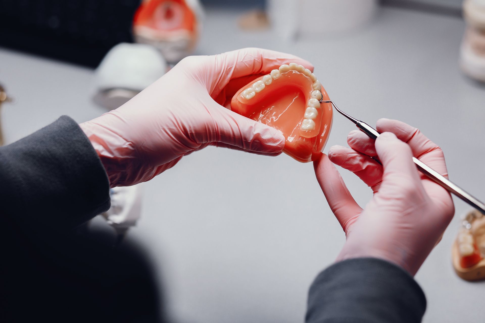 Hands in pink gloves hold a dental model, using a tool to adjust the teeth, on a white surface.