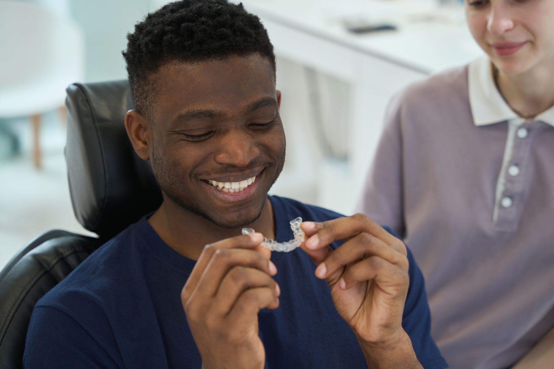 Man smiling, holding clear dental aligner, in a dentist's office. Woman nearby.