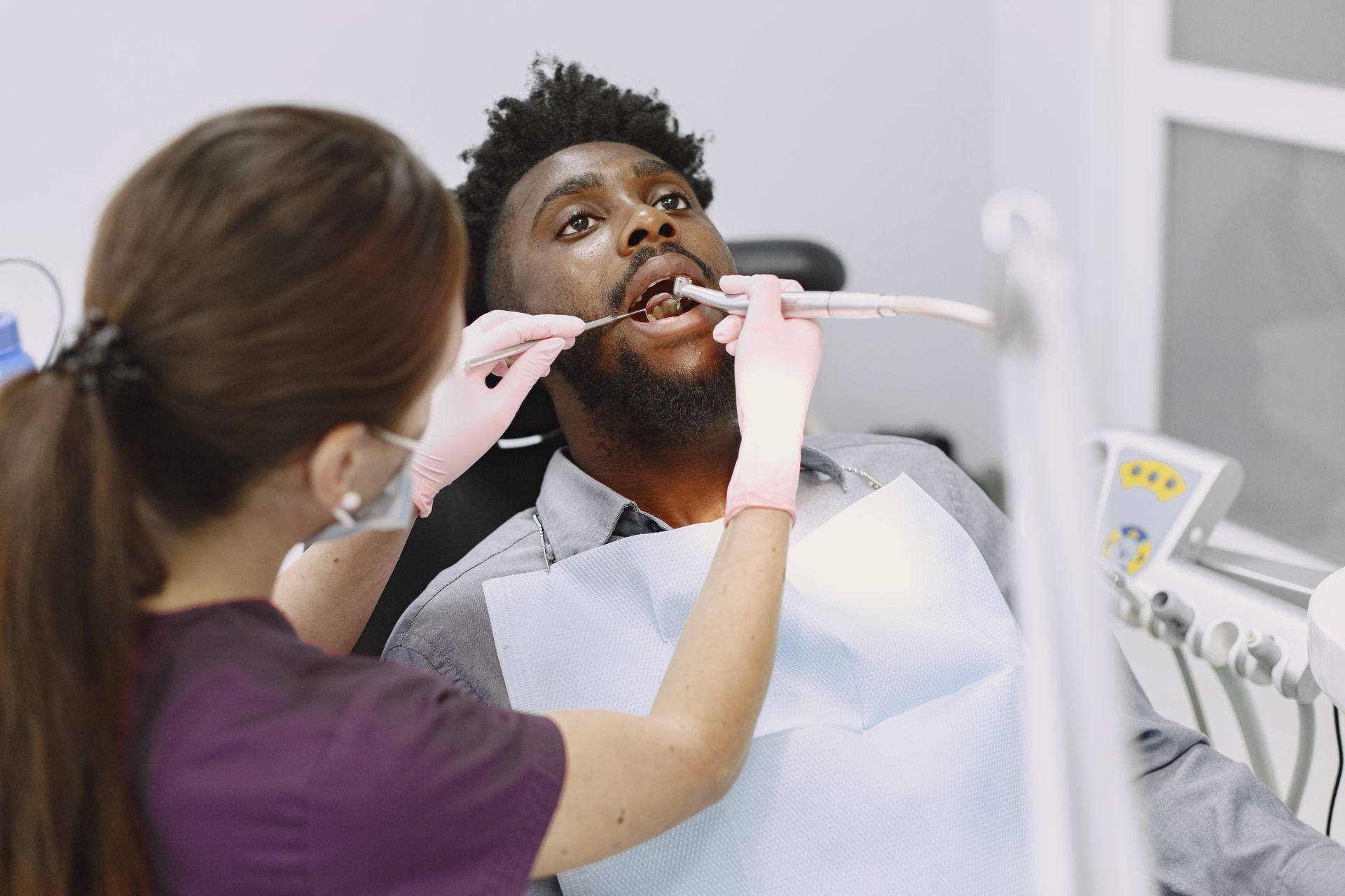 Dentist examining patient's mouth with tools; patient looks up. Dental office setting.