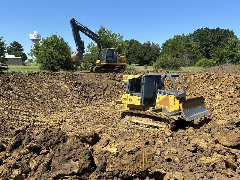 Yellow bulldozer on dirt site with excavator in background under a clear blue sky