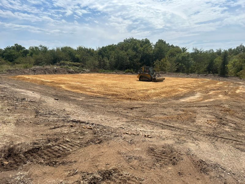 Skid steer grading a cleared dirt lot with trees in the background under a blue sky