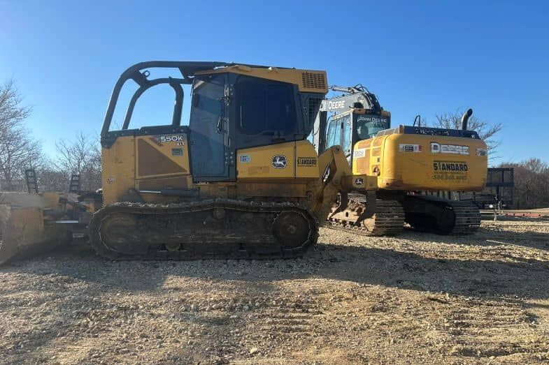 A yellow crawler tractor and an excavator parked side-by-side on a gravel lot under a clear blue sky.