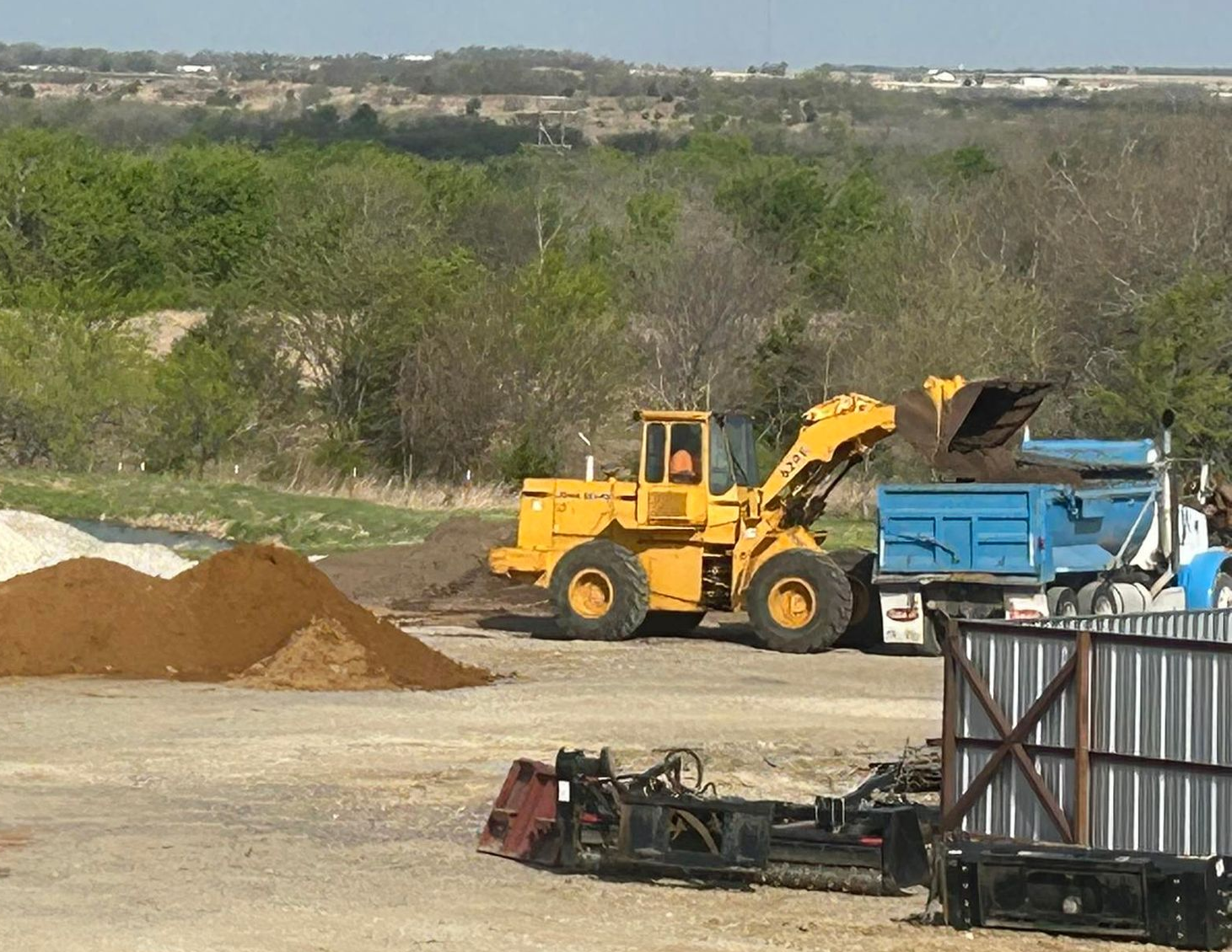 A yellow front-end loader dumps dirt into a blue dump truck at an outdoor construction or landscape supply site.