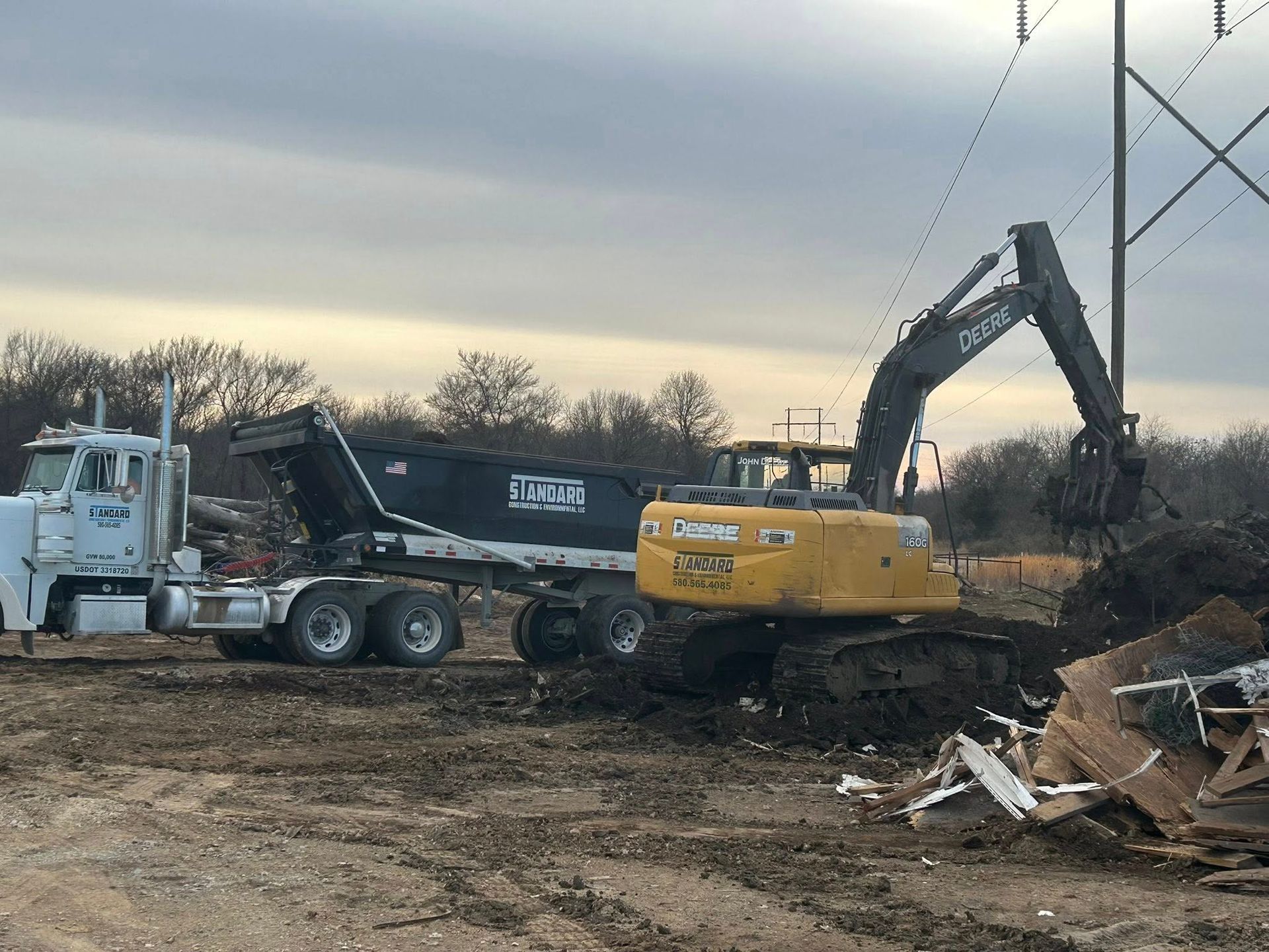 A yellow John Deere excavator loading demolition debris into a black dump truck under a cloudy sky.