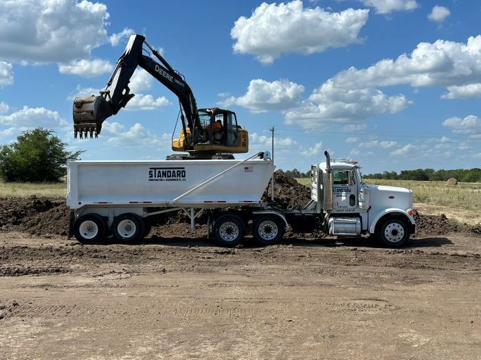 Excavator loading dirt into a white dump truck in a dirt lot under a blue sky.