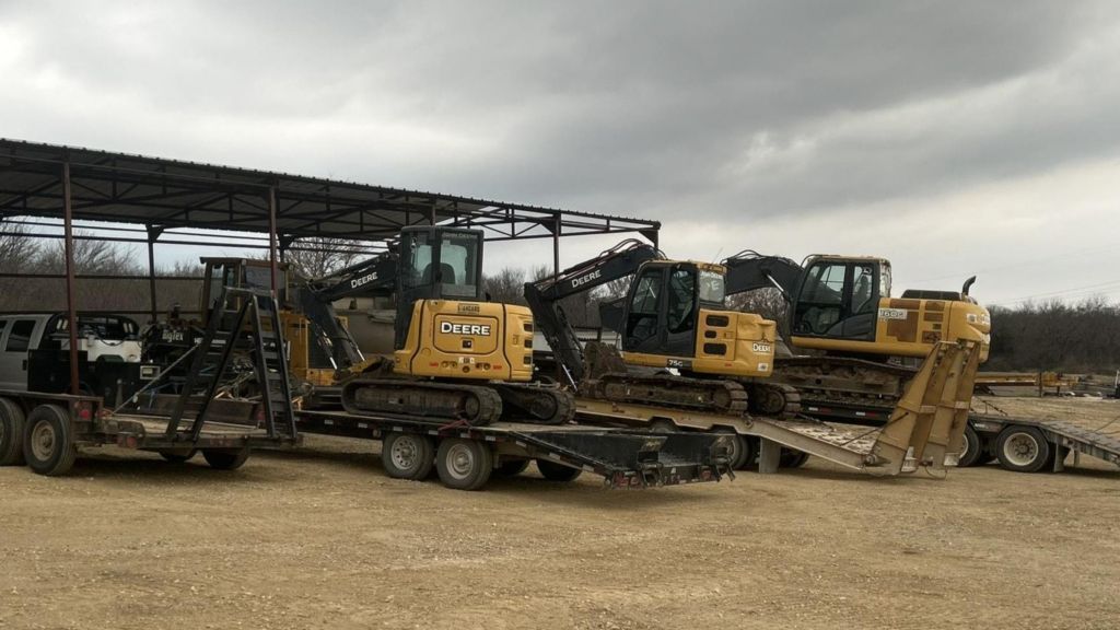 Several yellow excavators loaded on flatbed trailers under a cloudy sky in a gravel yard