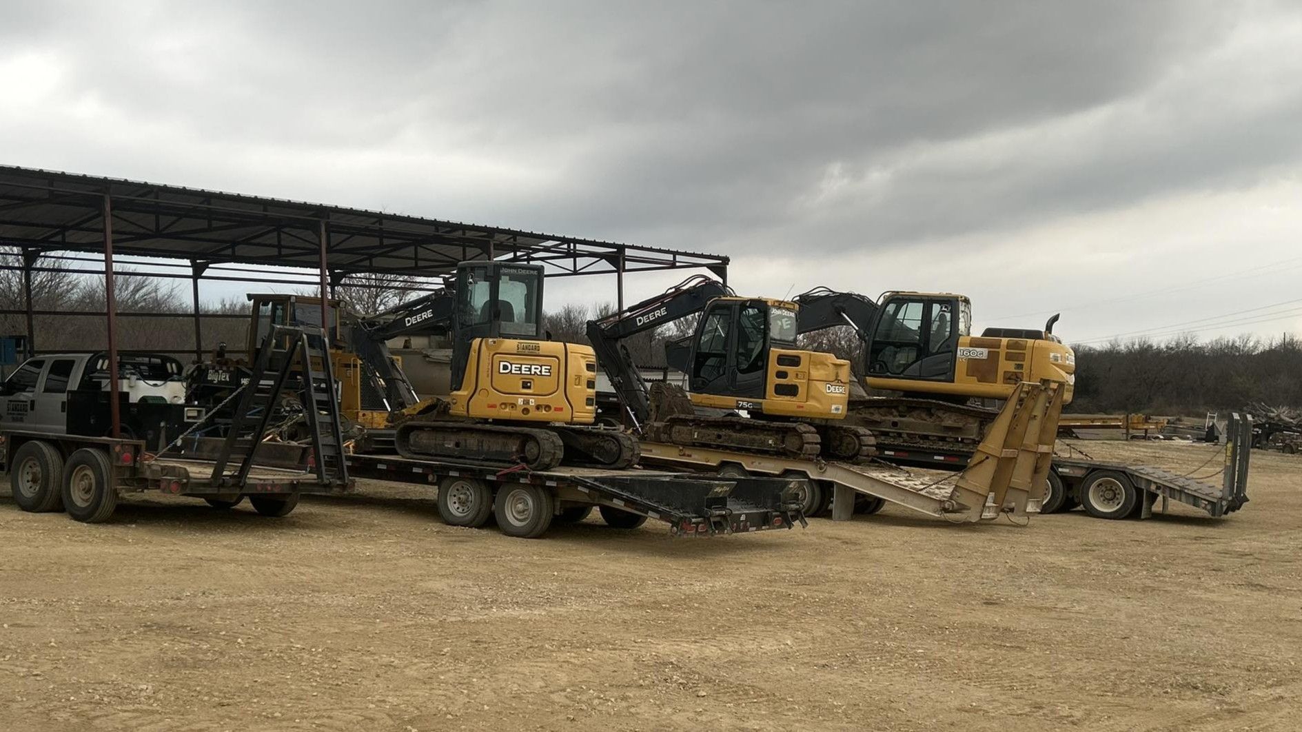 Flatbed trailers loaded with yellow construction equipment at a dusty outdoor site under a cloudy sky