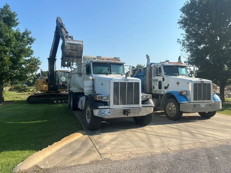 Two white dump trucks parked on a sunny driveway beside a tree-lined grassy area.
