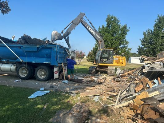 Excavator loading debris into a blue dump truck at a demolition site with scattered rubble and trees.