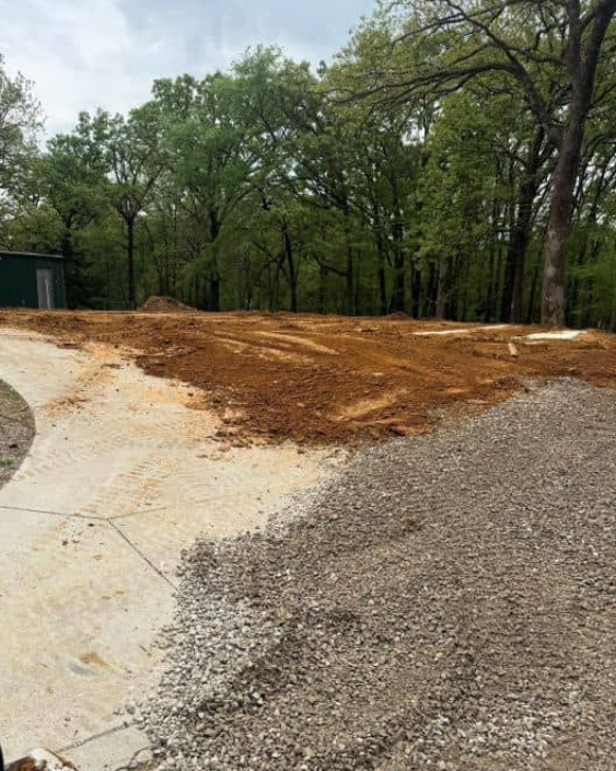 Gravel driveway under construction beside a wooded area, with fresh dirt spread across the path.