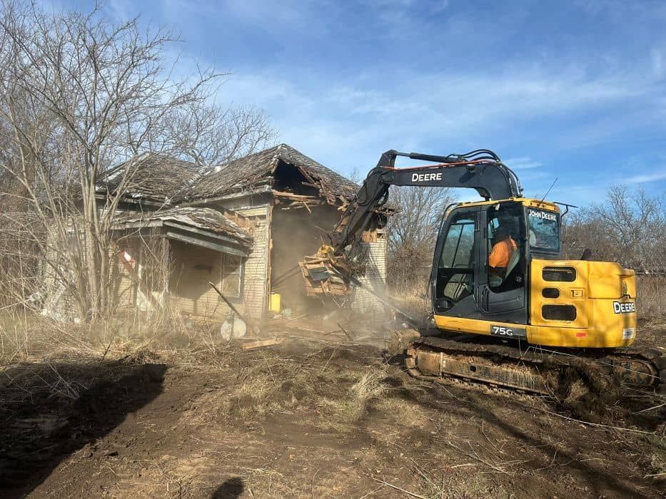 Yellow excavator demolishing a damaged house amid dust and bare trees