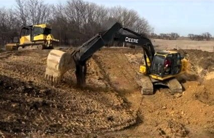A yellow John Deere excavator digs into a dirt embankment on a rural construction site with a bulldozer in the distance.